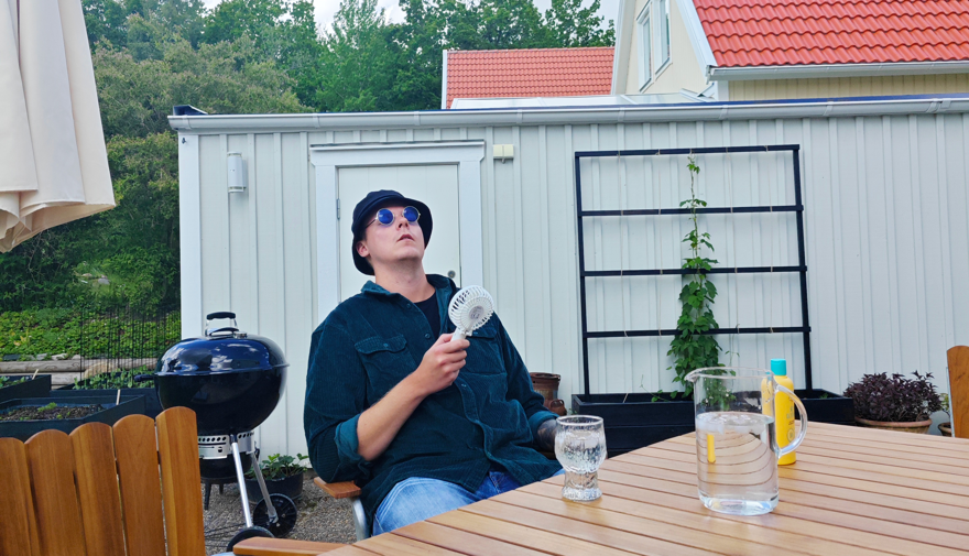 En man sitter utomhus på en stol med en fläkt i handen.