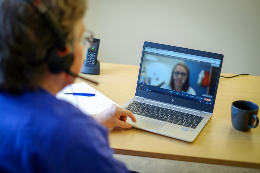 En rygg på en kvinna som sitter med headset framför en dator. På datorn har hon ett möte med en annan kvinna.