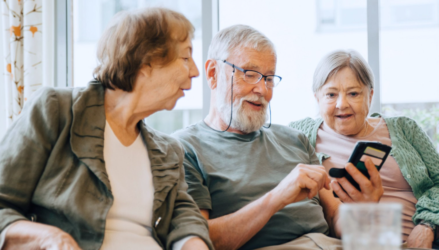 Två äldre kvinnor sitter bredvid en äldre man i en soffa. Mannen håller i en mobiltelefon. Samtliga kollar ner i telefonen och ser glada ut.